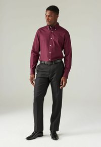 Cămașă burgundă cu guler, pantaloni negri de costum și pantofi negri. Cămașa are un logo discret brodat pe piept.