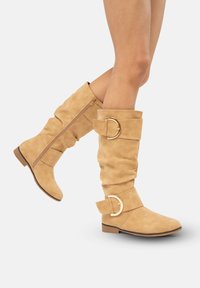 Botas de ante color beige hasta la rodilla con un diseño ajustado, que cuentan con un cierre lateral, grandes adornos de hebilla y una suela plana para mayor comodidad.