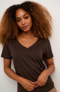 Camiseta de algodón marrón con cuello en V y mangas cortas, textura suave, diseño ajustado, llevada por una persona con cabello rizado, manos entrelazadas al frente.