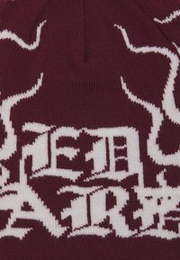 Bordově červená pletená čepice s bílými, složitými textovými vzory. Měkká textura, zaoblený vršek a žebrovaný okraj.