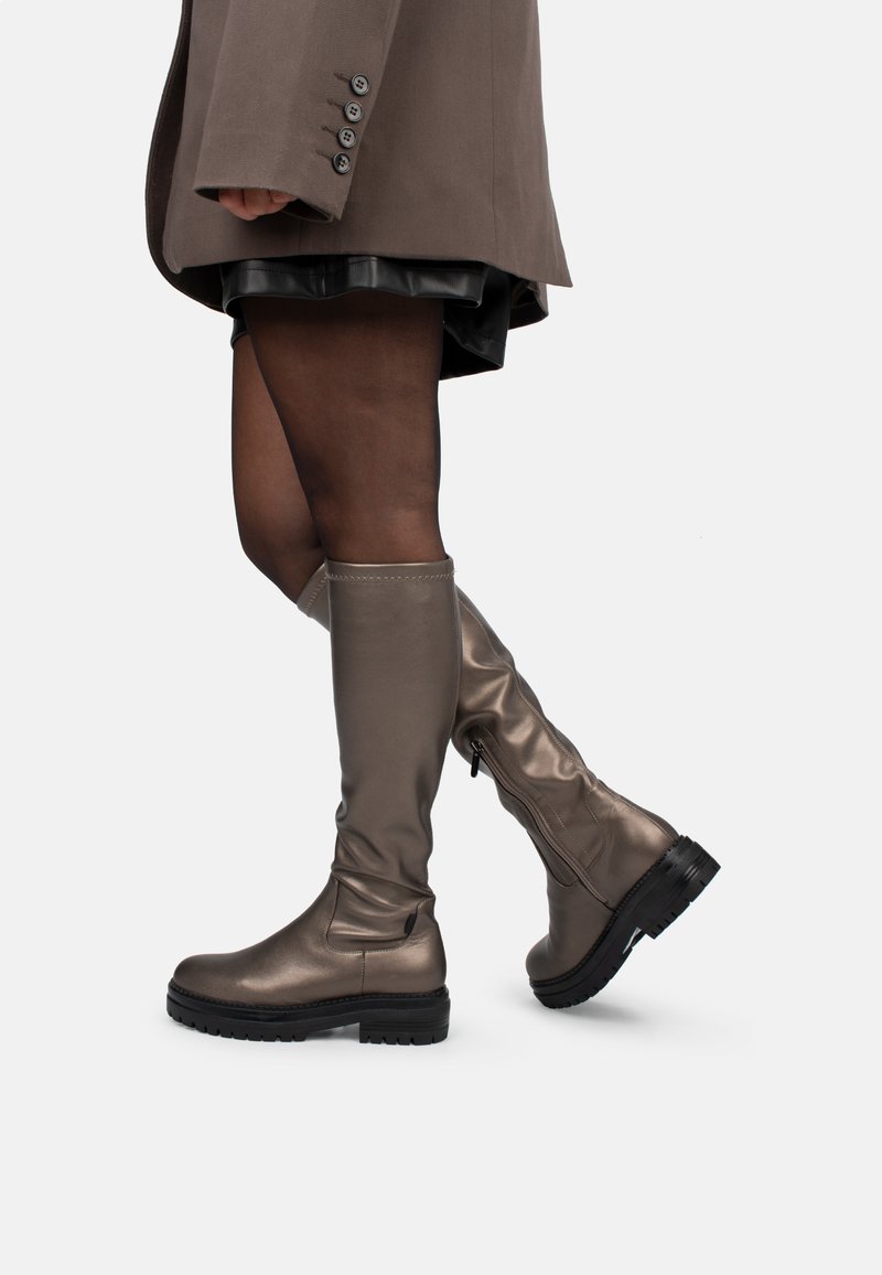 Botas de bronce metálico hasta la rodilla con un acabado suave, suela texturizada negra y detalle de cremallera lateral. Combinadas con una falda oscura y un abrigo.