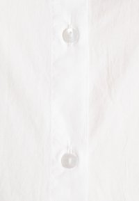 Camisa de algodão branca em close-up, apresentando uma pala de botões vertical com botões redondos transparentes e uma textura subtil, destacando o seu tecido suave.