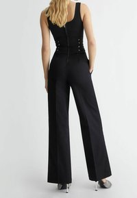 Mono negro con un cuerpo ajustado, pantalones de pierna ancha y cierre de cremallera en la parte posterior. Presenta detalles de cordones en la cintura y una textura de tela elegante.