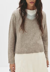 V-kaula, beige neulepusero, jossa on teksturoitu pinta, varustettuna ribatut hihansuut ja helma. Päällä vaaleanharmaa kerrospaita ja kerma-valkoinen hame.