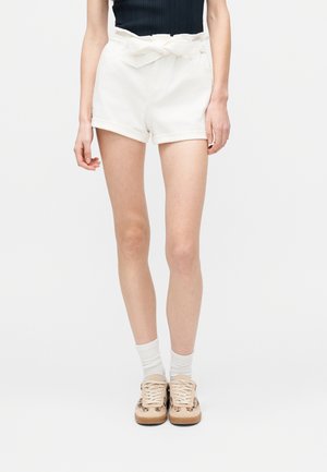 Γυναίκα που φοράει λευκά σορτς με ζώνη τύπου paperbag, μαύρο τοπ, λευκές κάλτσες και μπεζ αθλητικά παπούτσια με animal print λεπτομέρειες, στέκεται μπροστά σε λευκό φόντο.