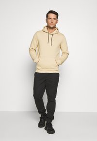 Beige katoenen hoodie met een voorzak en zwarte trekkoorden, gecombineerd met zwarte broek en zwarte sneakers. Gestructureerde pasvorm.