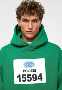 Groene hoodie met een groot wit voorpaneel dat "POLIZEI 15594" in het zwart toont. Heeft een dikke, gestructureerde stof en een diepe capuchon.