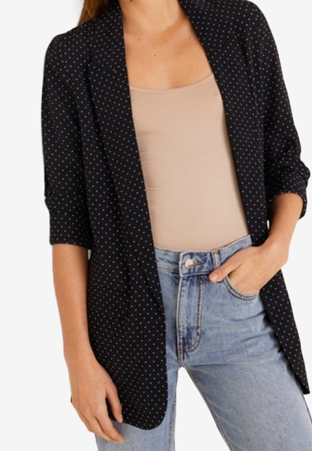 Schwarzer Blazer mit weißen Polka-Dots, drei Viertelärmel, aus weichem Stoff, mit offenem Vorderteil, kombiniert mit einem beigen Oberteil und hochgeschnittenen blauen Jeans.