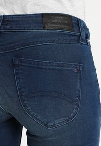 Jeans de mezclilla azul oscuro con una textura ligeramente desgastada, que cuentan con un bolsillo trasero con un diseño cosido y una etiqueta de marca de cuero negro.