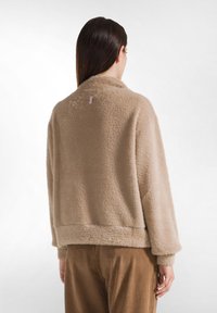 Suéter beige y suave con una textura blanda, cuello tipo solapa y diseño cropped. Presenta una etiqueta de marca visible en la parte posterior.