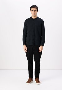Camisa negra de botones confeccionada en una tela texturizada, combinada con pantalones negros y zapatillas marrones. La camisa tiene un cuello redondo y mangas largas.