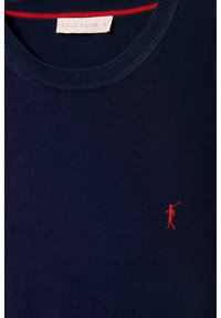 Námořnický modrý pletený svetr s kulatým výstřihem, který má červený detail uvnitř límce a malou červenou vyšívanou postavičku na hrudi.