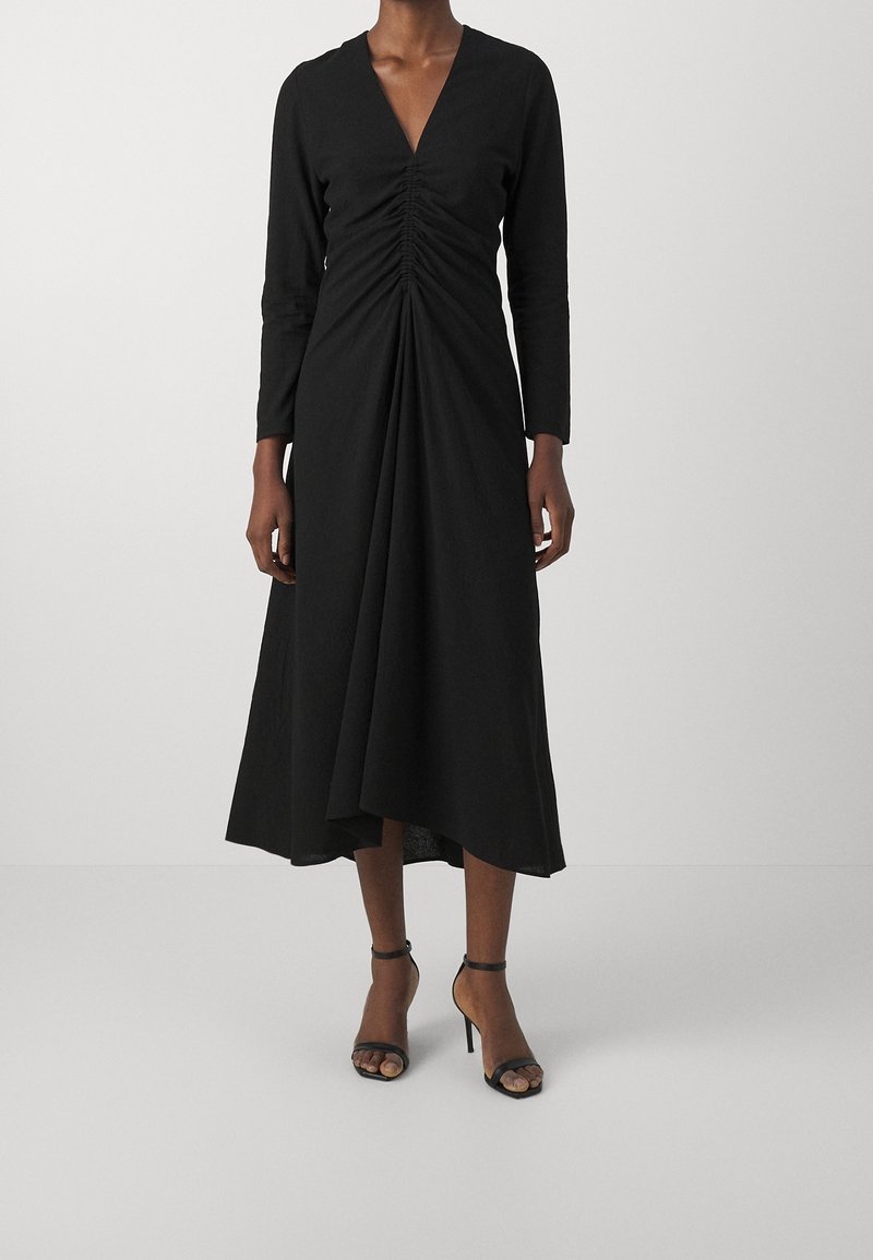 Femme portant une longue robe noire avec un détail froncé à l'avant, des manches longues, un décolleté en V et des sandales noires à talons hauts, debout sur un fond uni.