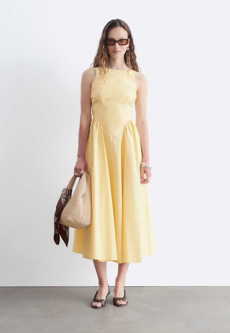 Femme portant une robe midi jaune sans manches, des lunettes de soleil foncées, tenant un sac à main tressé avec une écharpe marron, et portant des sandales marron.