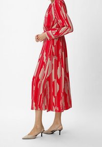 Femme portant une robe midi à manches longues rayée rouge et beige, associée à des chaussures à talons kitten beige avec bride au talon, se tenant de côté.