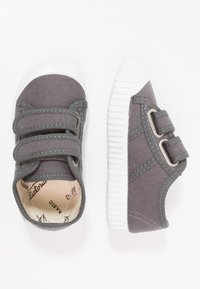 Sneakers in tela grigia con due cinturini in Velcro, suola in gomma bianca e dettagli di cucitura; interno morbido e testurizzato con etichetta del brand.