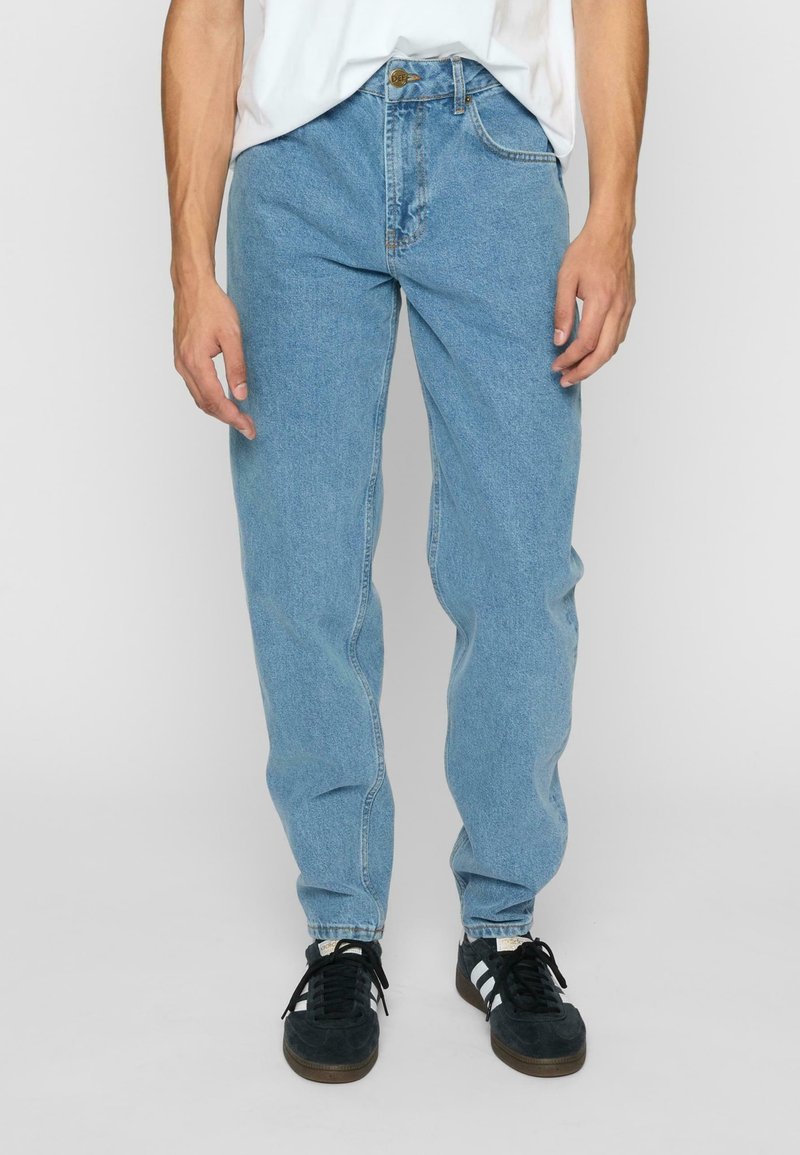 Homme portant un jean en denim bleu clair, une chemise blanche et des baskets noires avec des rayures blanches, debout devant un fond uni.