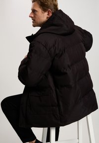 Czarna kurtka puchowa z kapturem, z pikowaną strukturą, dużymi kieszeniami z przodu oraz gładkim wykończeniem materiału. Model siedzi na stołku.