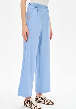 Jasnoniebieskie spodnie o szerokich nogawkach z guzikami w talii, noszone przez stojącą osobę, w połączeniu z białymi i brązowymi sportowymi butami w cętki.