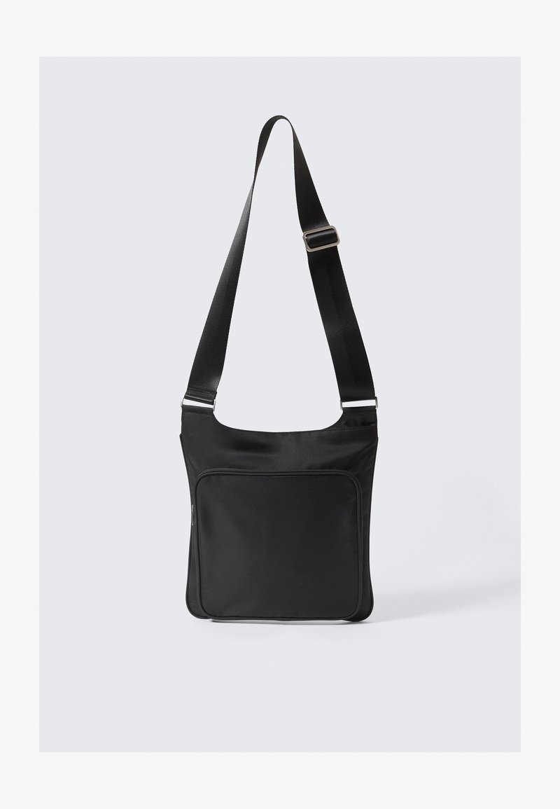 Bolsa de hombro negra de nailon con un diseño plano, correa ajustable y bolsillo con cremallera en el frontal. Presenta una textura suave y un estilo minimalista.