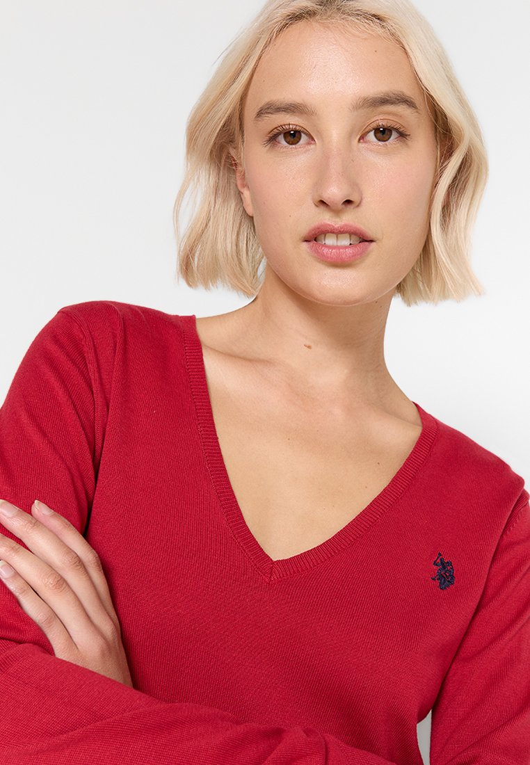 Junge Frau mit kurzen blonden Haaren, die einen roten V-Ausschnitt-Pullover mit einem kleinen gestickten marineblauen Logo auf der Brust trägt.
