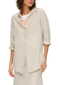 Beige Leinen-Hemdbluse mit einer einzigen Brusttasche, hochgekrempelten Ärmeln und einem abgerundeten Saum. Getragen über einem weißen T-Shirt mit oranger Schrift.