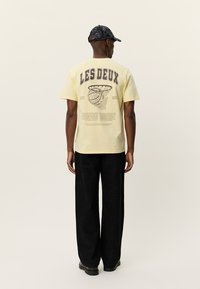 Κίτρινη βαμβακερή μπλούζα με την επιγραφή "LES DEUX" στην πλάτη, με γραφικό σχέδιο και λεπτομέρειες κειμένου. Συνδυασμένη με μαύρο παντελόνι με μοτίβα.