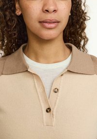 Bežani pleteni polo majica s kontrastnim svijetlosmešanim ovratnikom, s dva dugmeta i mekim, teksturiranim završetkom. Nosena preko svijetle majice.