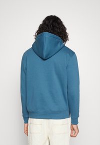 Persona con capelli ricci che indossa una felpa con cappuccio blu e pantaloni testo panna, in piedi di spalle su uno sfondo bianco semplice.