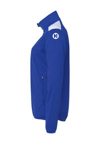 Blå atletisk jakke med høj krave, hvide detaljer på skulderen, lynlås-lomme og et cirkulært logo på ærmet. Blød stoftekstur.