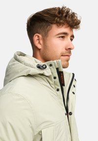 Lys beige jakke med hette, med en frontglidelås, knapper og et oransje merke. Glatt stoff og standard passform.