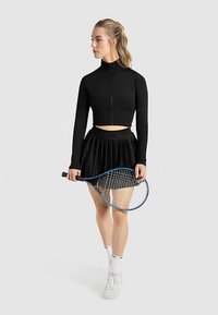 Schwarzes, kurzes Langarmshirt und plissierter schwarzer Rock. Das Model hält einen Tennisschläger, trägt weiße Socken und Schuhe. Minimalistisches Design.