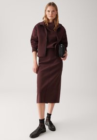 Bordowa, oversize kurtka na dopasowanej bluzce i midi spódnicy. Model nosi czarne loafersy z przezroczystymi skarpetkami do kostek. Gładka tekstura materiału.