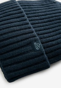Écharpe en tricot bleu marine foncé avec texture côtelée, présentant une petite étiquette de marque avec un design géométrique. Matière douce et chaude.