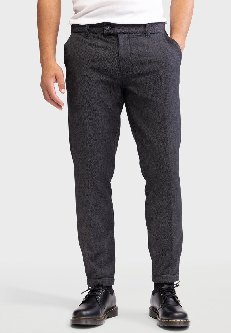 Homme portant un pantalon gris foncé ajusté avec revers, des chaussures noires à lacets, et une chemise blanche rentrée, debout sur un fond clair.