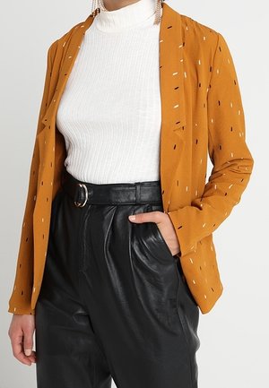 Frau trägt einen senffarbenen Blazer mit kleinen schwarzen und weißen Strichen, weißen gerippten Rollkragenpullover, hochtaillierte schwarze Lederhosen mit Gürtel, Hand in der Tasche.