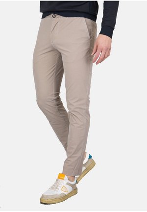 Homme portant un pantalon beige slim, des baskets blanches avec des accents orange et bleu, et un haut à manches longues foncé, debout avec une jambe légèrement pliée.