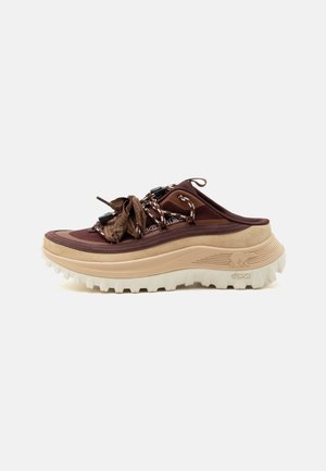 Καφέ και μπεζ αθλητικό παπούτσι τύπου slip-on με ανθεκτική λευκή σόλα, ρυθμιζόμενα κορδόνια τύπου bungee και θηλιά στον αστράγαλο, σε λευκό φόντο.
