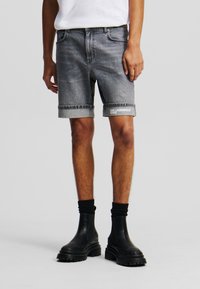 Pantalones cortos de mezclilla gris con dobladillos enrollados, que presentan un logotipo en una pierna. Usados con botas negras hasta el tobillo y calcetines. Corte sencillo, diseño clásico.