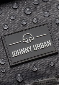 Svart texturerad tyg med vattendroppar; har en rektangulär gummimärket med upphöjd vit text "JOHNNY URBAN" och en logotyp i mitten.