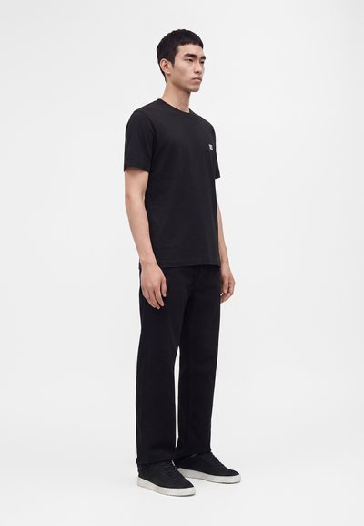 Homme debout portant un t-shirt noir uni, un pantalon noir et des baskets noires avec semelles blanches sur fond blanc.