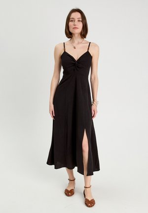 Frau in einem schwarzen ärmellosen Midikleid mit Vorderschlitz, trägt braune Schuhe mit Knöchelriemen und eine Halskette, vor einem schlichten hellen Hintergrund.