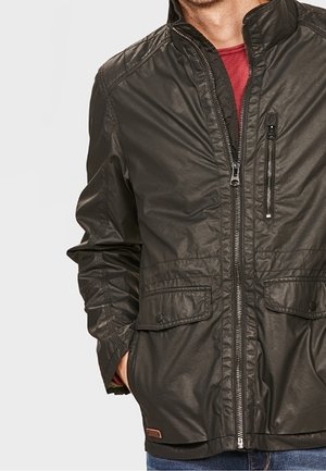 Mann trägt eine dunkelbraune Reißverschlussjacke mit Klapptaschen und einer Reißverschlusstasche auf der Brust über einem roten Hemd, Hände in den Taschen.