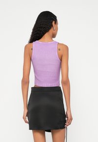 Žena s dlhými kučeravými vlasmi, oblečená v bezrukávovom fialovom crop tope a čiernej mini sukni, stojaca chrbtom k nám na jednoduchom pozadí.