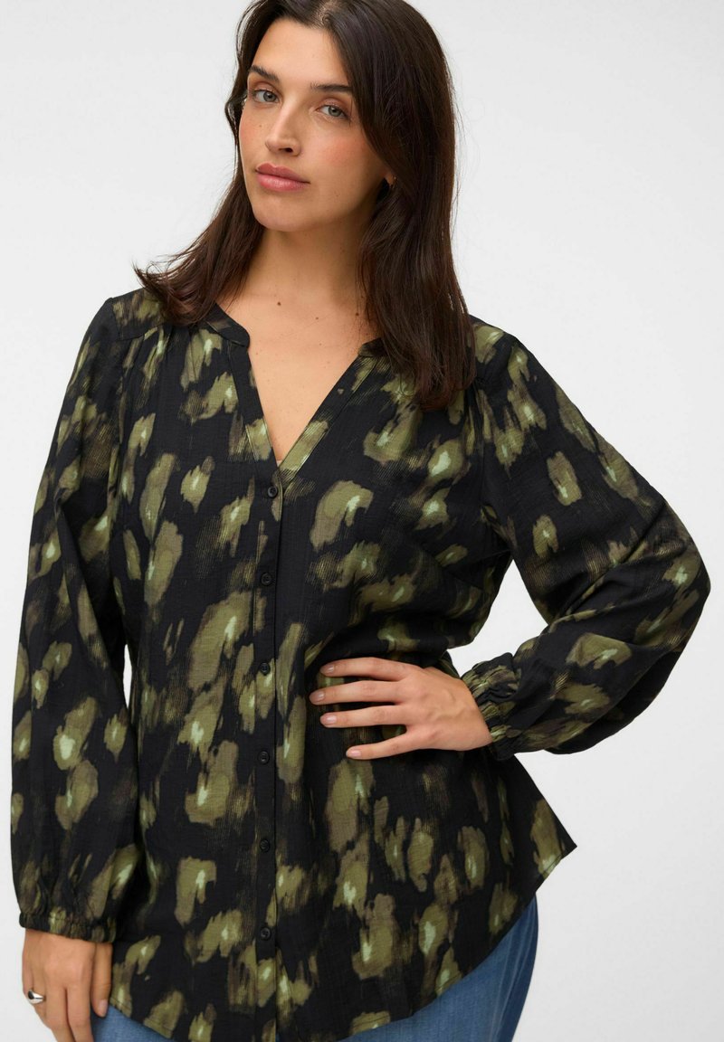 Femme aux cheveux foncés mi-longs portant un chemisier noir boutonné avec des motifs abstraits verts et un jean bleu, posant avec une main sur la hanche.