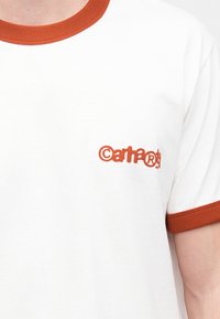 Λευκό βαμβακερό μπλουζάκι με πορτοκαλί ριμπ γιακά και μανίκια. Φέρει το κείμενο "©artha®" κεντημένο σε πορτοκαλί στην αριστερή περιοχή του στήθους.