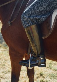 Stivali da equitazione in gomma nera con finitura lucida, caratterizzati da un accento argentato e fibbia, abbinati a pantaloni da equitazione grigi con motivo, su un cavallo.