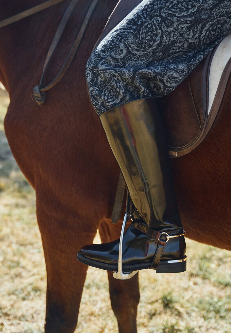 Stivali da equitazione in gomma nera con finitura lucida, caratterizzati da un accento argentato e fibbia, abbinati a pantaloni da equitazione grigi con motivo, su un cavallo.