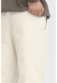 Pantalones de algodón color crema con un corte relajado, que cuentan con dos bolsillos traseros y un bolsillo lateral. La tela parece suave y ligera.