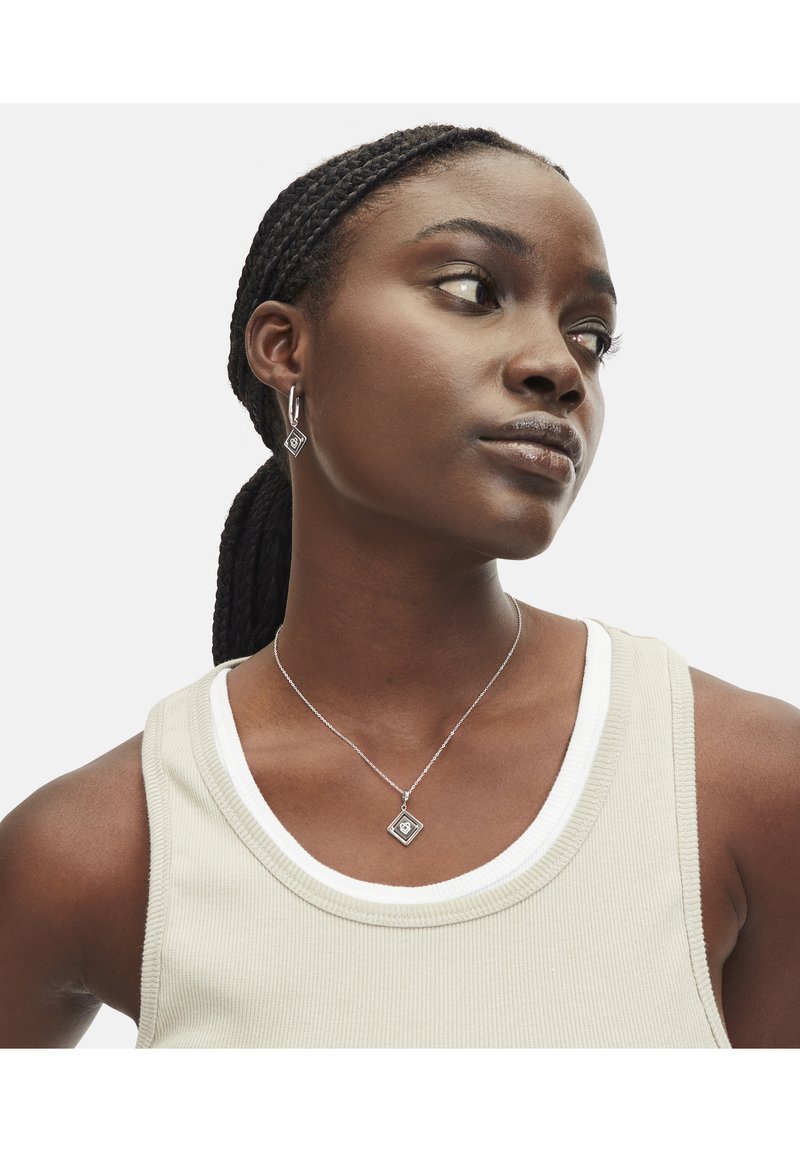 Zilveren sieradenset met een diamanten hanger en bijpassende oorbellen. Draagt over een geribbelde, lichtgekleurde tanktop.